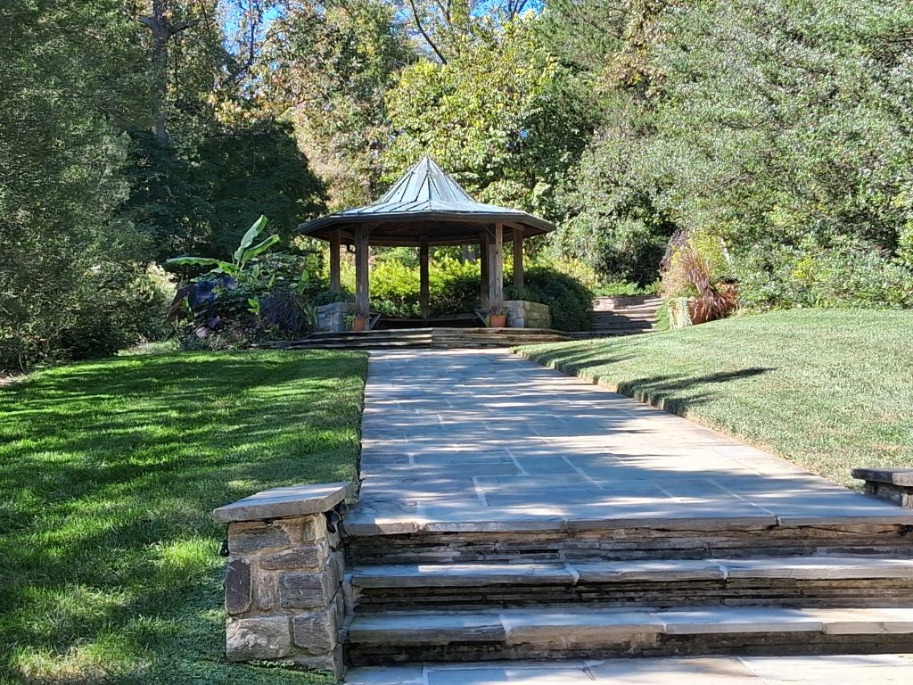 Stone steps and a paved path lead to a shaded garden pavilion surrounded by grass and trees at Brookside Gardens.