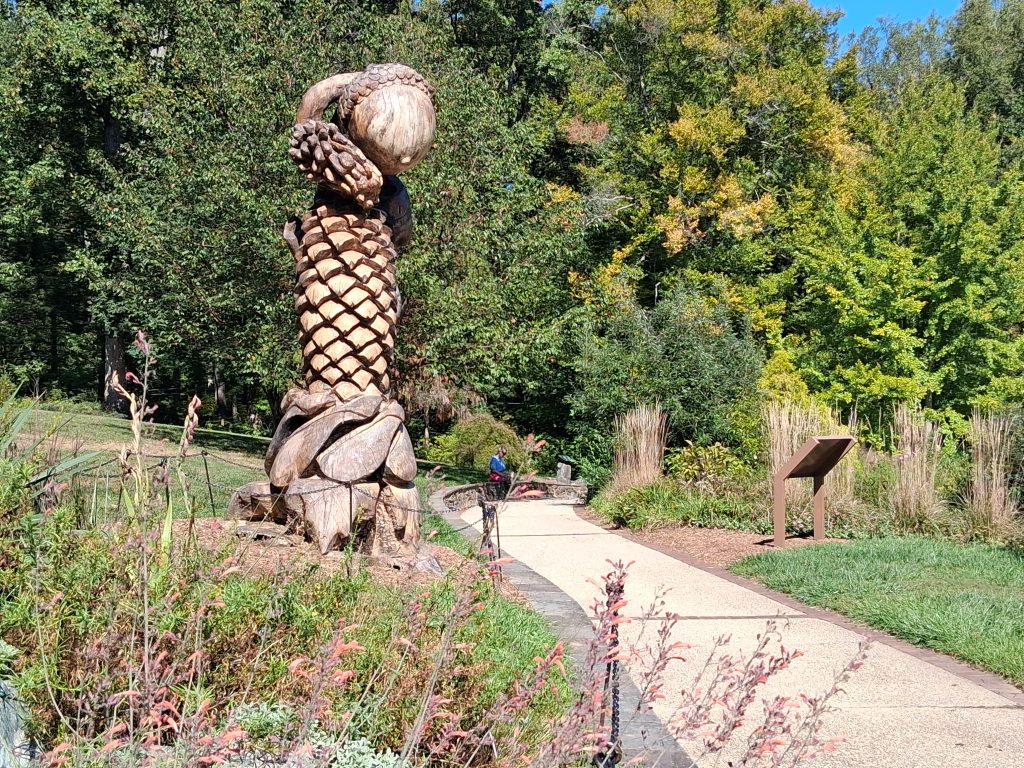 Wooden acorn and pine-cone sculpture beside a curving path, planting beds, and an information sign at Brookside Gardens.